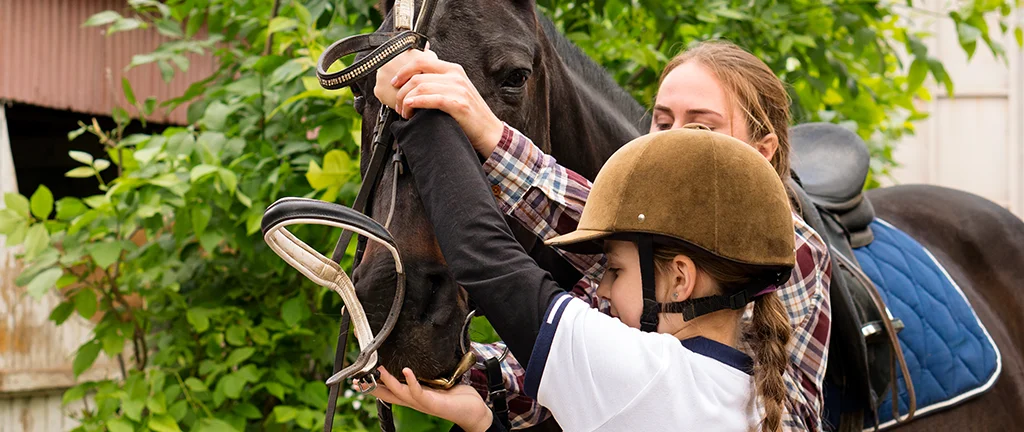 Girl and instructor fitting a bridle on a brown horse outdoors.