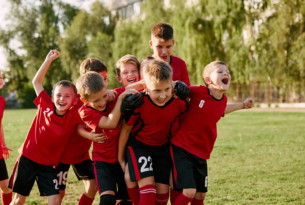 Joyful students in team tricots playing soccer on a grassy soccer field.